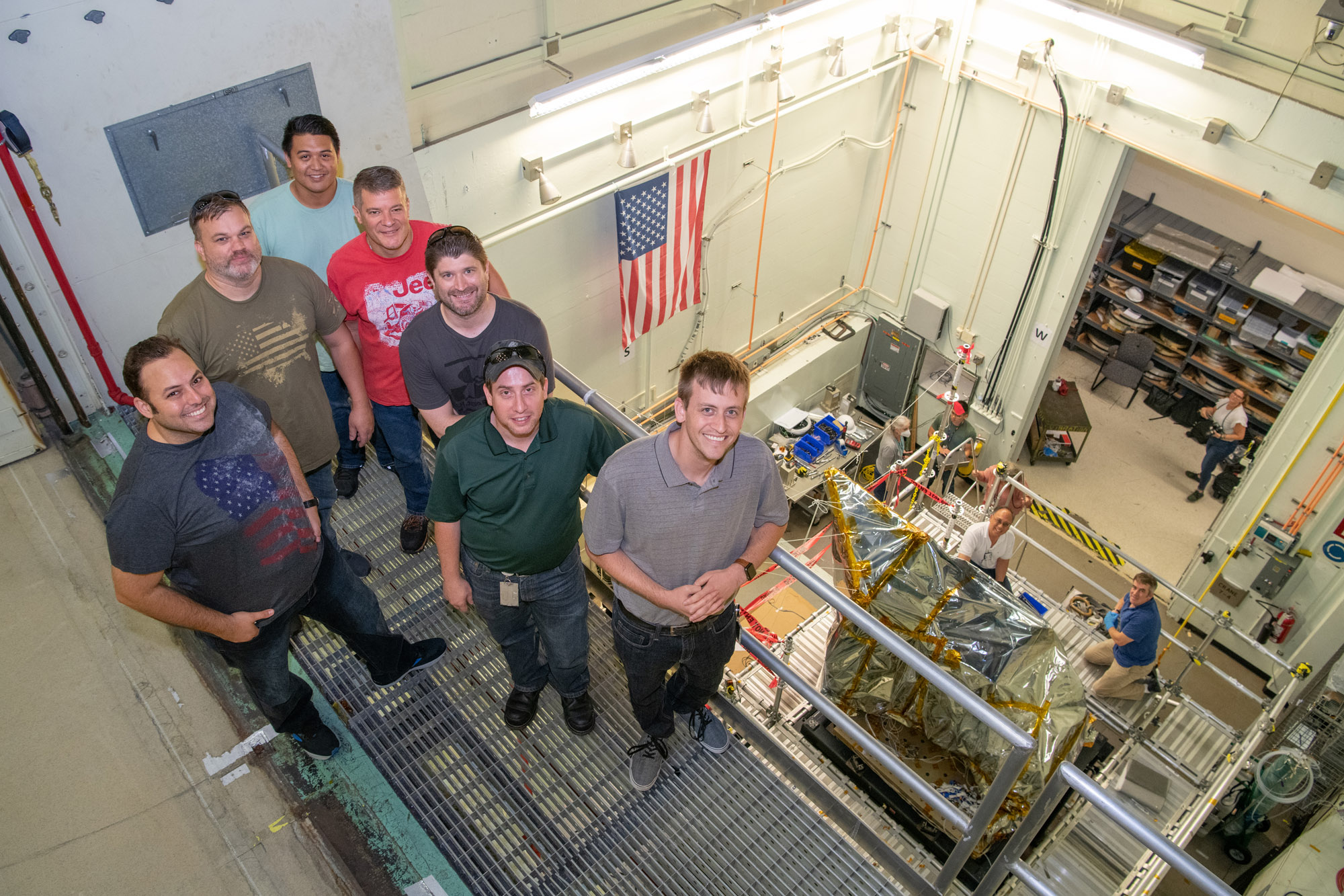 The Ocean Color Instrument mechanical team stands on the balcony overlooking the bagged instrument after successfully installing it onto the X axis (vertical) vibration shaker table.