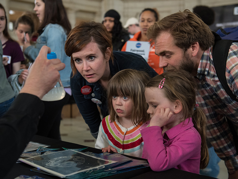 A family checks out a vial containing a culture of <em>Emiliana huxleyi</em>, a phytoplankton that plays an important role in the global carbon cycle. Credit: Aubrey Gemignani (NASA)